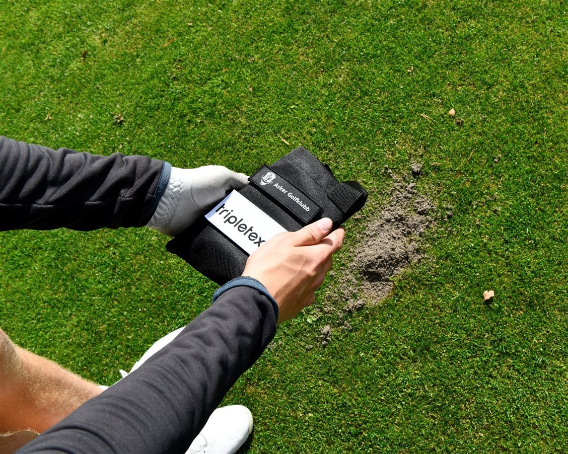 Golfspiller holder en Tripletex-merket sandpose fra Asker Golfklubb over et slagmerke